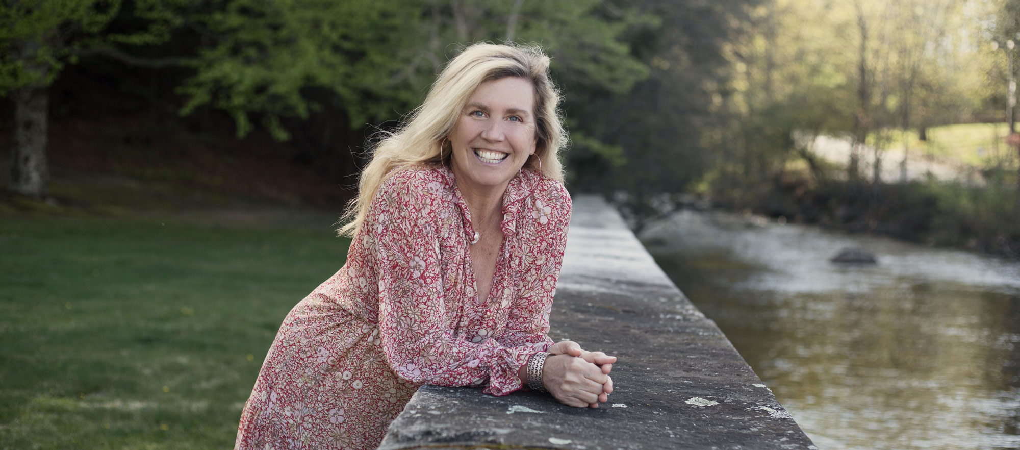 Kasey Mathews smiling wide while leaning over the edge of a stone barrier. On the other side of the stone barrier is a river. Kasey is wearing a red floral dress with long ruffled sleeves and a silver bracelet